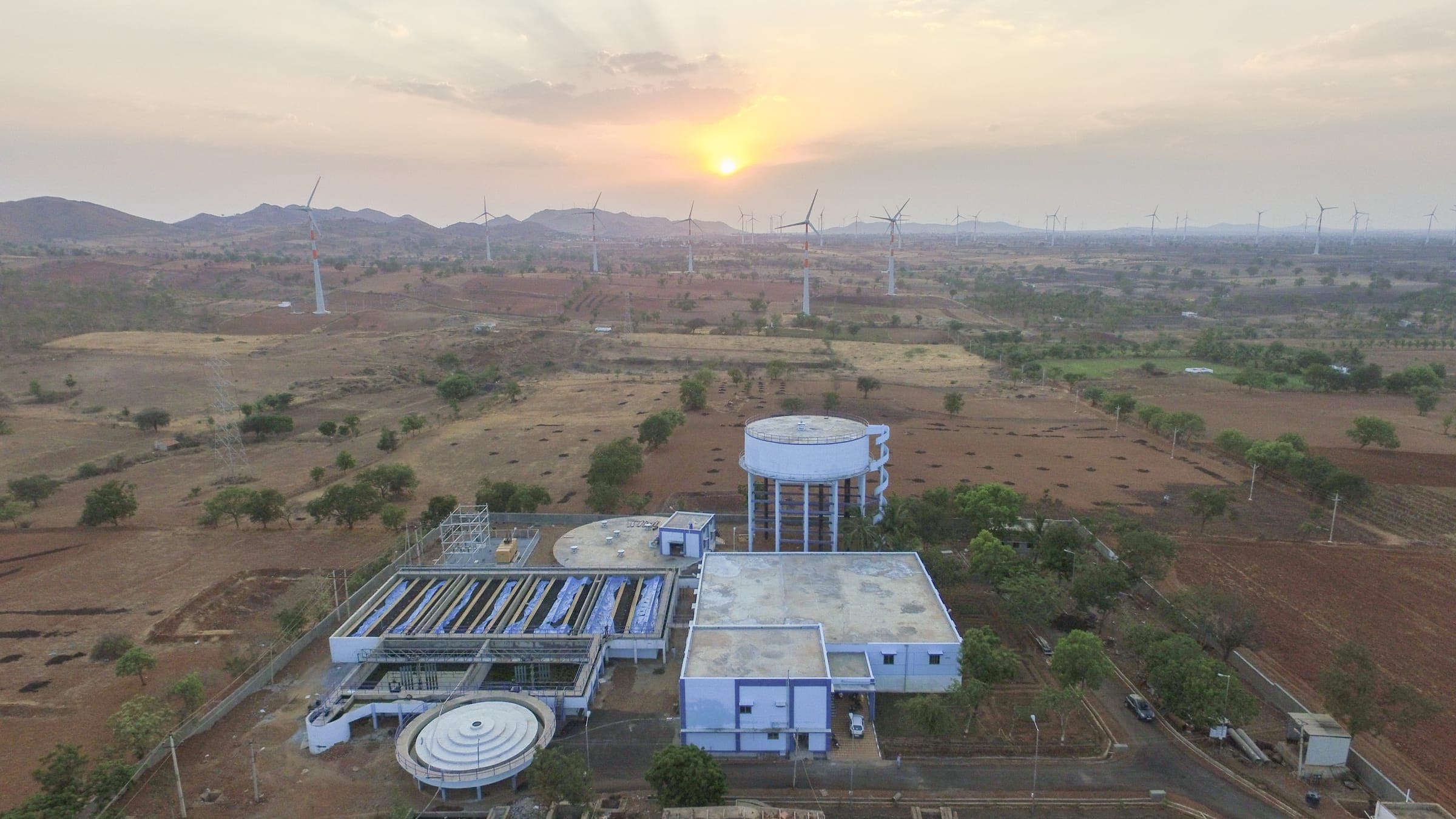 Aerial view at sunset of a Gondwana water treatment plant — rectangular filter beds, a circular clarifier, an elevated clearwater tank, and the control building against rolling hills and a wind farm